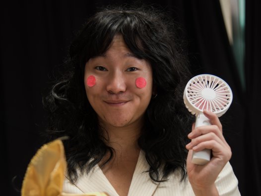 Ein asiatischer gelesener Mensch mit einer schwarzen Langhaar-Perücke und roten Papierpunkten auf den Backen hält einen kleinen Handventilator hoch und lächelt verschmitzt. | © ©Gedvilė Tamošiūnaitė