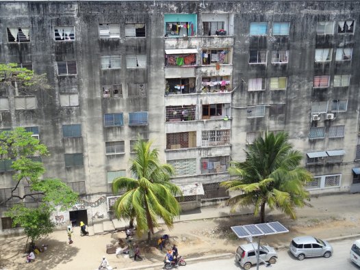 Die Fassade eines Plattenbaus in Sansibsar und ein Ausschnitt der davorliegenden Straße mit Autos und Palmen | © Sarah-Marleen Methner